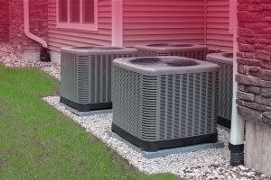 A photograph of four grey, metal AC condensing units, part of a multi-unit residential HVAC system, are neatly arranged in a row on concrete pads within a gravel bed against the side of a house featuring light-colored siding and stone veneer accents.