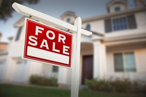A prominent red and white "For Sale" sign on a white post with a modern suburban home blurred in the background.