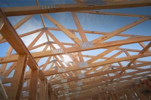 An upward view of wooden roof trusses and framing under a clear blue sky with sunlight shining through.
