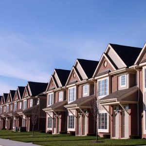 Row of multi-story brick and siding suburban townhomes with dark shingle roofs and prominent gables under a blue sky.