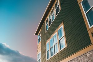 Low-angle exterior view of a multi-story modern residential building with dark green siding, white-framed windows, and a soft sunset sky.